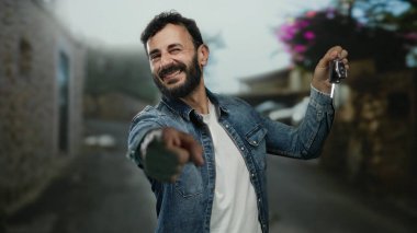 İspanyol adam elinde arabanın anahtarını tutuyor. Kot ceket giyiyor. Heyecanlı ve mutlu görünüyor. Kırsal binaların ve çiçeklerin bulanık arka planına sahip anahtarları işaret ediyor..