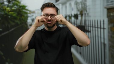 Sakallı ve gözlüklü genç adam şehir sokaklarında dikiliyor. Etrafı kentsel binalarla çevrili.