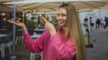 Dışarıdaki bir restoranda pembe bir gömlek giyerken terasta gülümseyen ve işaret eden bir kadın, canlı bir yemek sahnesi ve sıradan bir ortam sergiliyor..