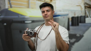 Şehir sokağındaki İspanyol asıllı genç adam elinde antika bir kamera tutarken parmaklarıyla şakacı jestler yapıyor ve canlı şehir sahnesini yakalıyor..
