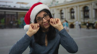 Gözlüklü kadın kırmızı Noel Baba şapkası takıyor ve şehir binasının önünde yumruklarını yanaklarına bastırıyor; bayram nostaljisi üzüntüsü pişmanlığı.