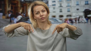 Arka planda bulanık bina cepheleriyle şehir caddesinde her iki işaret parmağı aşağı doğru olan kadın dikkat istiyor.