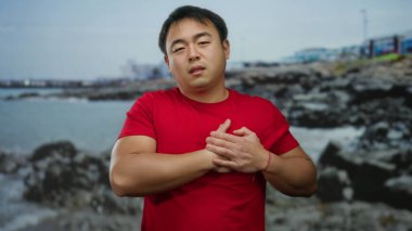 Bulutlu bir günde deniz kenarında göğsü ağrıyan, kayalık bir geçmişi olan, endişeli ve düşünceli görünen kırmızı gömlekli genç bir adam..