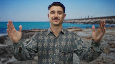 Genç İspanyol adam açık mavi bir deniz ve gökyüzü ile güneşli bir sahil tarafında, açık bir el hareketi yapıyor, desenli bir gömlek giyiyor, neşeli ve rahat bir açık hava atmosferi taşıyor..