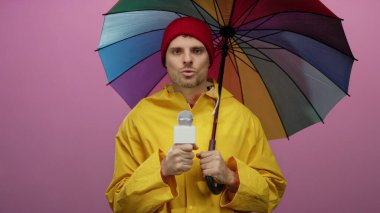 Sarı yağmurluklu adam mikrofon tutuyor. Pembe arka plana karşı çok renkli şemsiye altında duruyor.
