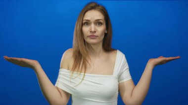 Beyaz bluzlu çıplak omuzlu kadın avuçlarını yukarı doğru tutar mavi stüdyoda şaşkın bir ifadeyle; belirsizlik.
