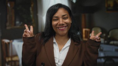 Kahverengi ceket ve beyaz gömlek giyerken restoranda gülümseyen ve barış işareti yapan bir kadın..