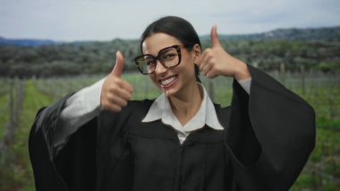 Yargıç üniforması içinde gülümseyen bir kadın, manzaralı bir parkta başparmak kaldırıyor, mutluluk ve profesyonelliği canlı yeşil bir arka planda somutlaştırıyor..