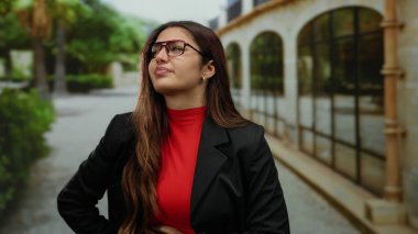 Yeşil ve kemerli mimariyle çevrili kentsel bir ortamda, gözlüklü ve kırmızı balıkçı yakalı bir yaka giyen bir kadın dışarıda acı dolu bir ifadeyle dikiliyor..