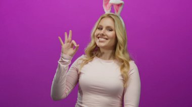 Tavşan kulağı takan ve parlak pembe arka plana karşı iyi bir hareket yapan, neşeli davranışlar sergileyen, sarışın ve genç görünümlü neşeli ifadeler sergileyen bir kadın..