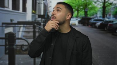 Elleri çenesinde genç İspanyol bir adam park halindeki arabalarla dolu bir şehir caddesinde yukarı bakıyor..