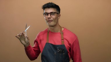 Gözlüklü ve önlüklü genç bir adam kahverengi bir arka plana karşı makas tutuyor ve berberlik mesleğini gösteriyor..