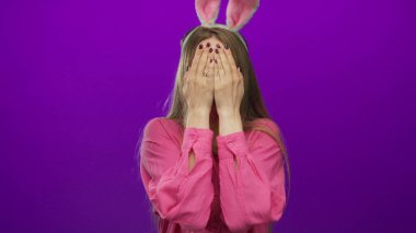 Pembe gömlekli, paskalya tavşanı kulaklı genç bir kadın mor arka planda şakacı yüzünü örtüyor..