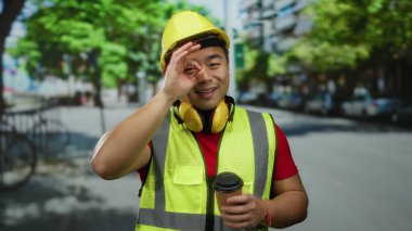 Güvenlik kaskı ve yelek giyen gülümseyen adam gündüz vakti elinde kahve tutuyordu.