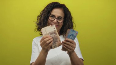 Orta yaşlı, gözlüklü, Meksika pezosu tutan İspanyol bir kadın izole edilmiş sarı bir duvar arkasına yaslanmış, neşeli bir ifadeyle para gösteriyor..