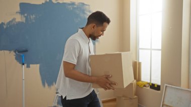 Yeni boyanmış oturma odasında kutu taşıyan genç bir adam, yeni bir daireye taşınmayı ve ev dekorasyonunu öneriyor, İspanyol kimliğini ve ev ortamını ön plana çıkarıyor..