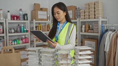 Yansıtıcı yelek giyen genç bir kadın bir hayır kurumunda çalışıyor. İçerideki malzemeleri düzenliyor. Üniforması ve tabletiyle gönüllü ruh ve verimliliği vurguluyor..