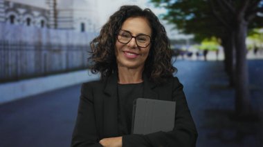 Orta yaşlı İspanyol kadın güvenle gülümsüyor şehir sokak ortamında elinde tabletle bulanık arka plan ağaçları ve binalarla..