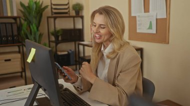 Masada akıllı telefon kullanarak modern dekor ve bitkilerle dolu profesyonel bir iş ortamında sevinç ve heyecanı ifade eden sarışın kadın, ofisinde kapalı yumrukla kutlama yapıyor.