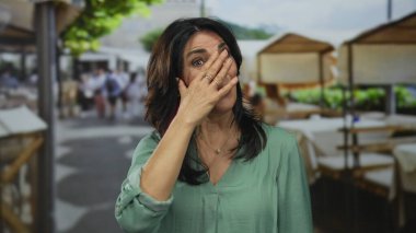 Yeşil bluzlu orta yaşlı bir kadın, parlak güneş ışığı altında terasta elini açık tutarak alnına dokunur..