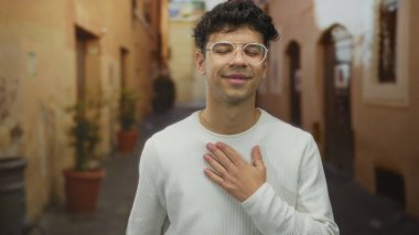 Dar kaldırım taşı caddesindeki yıpranmış binaların yanında gözlüklü ve beyaz kazaklı genç adam..