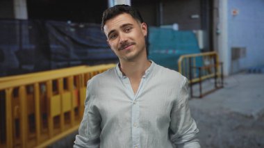 Bir inşaat alanında gülümseyen İspanyol genç adam, elleri şakacı bir şekilde kaldırılmış, arka planda inşaat engelleri olan açık renk bir gömlek giymiş..