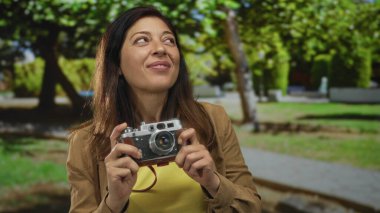 Güneşli bir ormanda iki eliyle klasik bir kamera tutan bir kadın, hafifçe gülümserken, huzur....