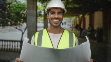 Şapkalı ve yansıtıcı yelekli genç İspanyol adam geniş bir plan tutuyor ve sokakta plan okuyor; profesyonel güven..