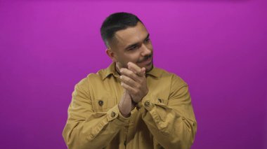 Kısa koyu renk saçlı, kahverengi ceketli, el çırpan bir adam parlak mor stüdyo duvarına bakarken, huzur içinde..