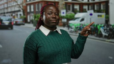 Şehir caddesinde gülümseyen bir kadın yeşil süveter ve örgü takmış neşeli bir ifade ve canlı bir çevre sergiliyor..