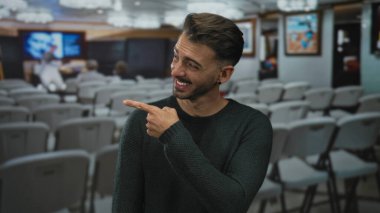 Yeşil kazak giyen bir adam binanın içindeki çerçeveli resme gülümseyerek parmak işareti yapıyor; merak neşe kaynağı..