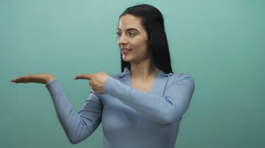 Mavi duvara karşı el ve baş parmaklarıyla el kol hareketi yapan, mavi bluz giyen, canlı bir iç mekanda pozitiflik ve iletişim kuran bir kadın..