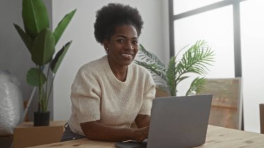 Modern oturma odasında dizüstü bilgisayar kullanırken gülümseyen bir kadın. Etrafı bitkiler ve kutular ile çevrili. Rahatlamış ve rahat bir atmosferi olan yeni bir ev ortamı öneriyor..