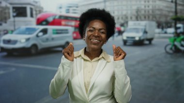 Kalabalık bir şehir caddesinde, arka planda kırmızı çift katlı otobüsleri olan beyaz bir spor ceket giymiş, güvenli bir şekilde gülümseyen bir kadın..