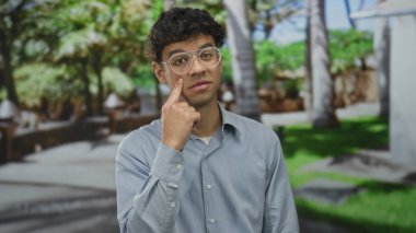 Gözlüklü ve açık mavi gömlekli bir adam, yeşil parkta bulanık banklar ve ağaçlarla parmak yanağa duruyor..