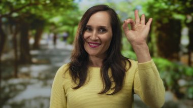 Sarı elbiseli kadın, yeşil yapraklı bir parkta kendinden emin ve dışa dönük bir şekilde iyi bir jest yapıyor. Çevresinden memnun olduğunu gösteriyor..