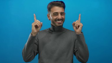 Mavi stüdyo ortamında gri balıkçı yakalı adam gülümsüyor ve her iki işaret parmağı yukarı doğru; iyimserlik umut pozitifliği.