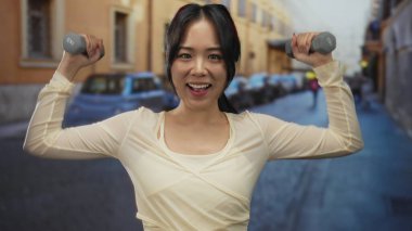 Sokakta halterleri kaldıran genç bir kadın neşeli bir ifadeyle, arka planda araçlarla şehir ortamında zindelik ve güç gösterisi yapıyor..