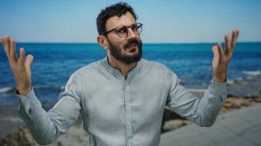 Sakallı ve gözlüklü İspanyol bir adam deniz kenarında mavi bir deniz ve açık bir gökyüzüne karşı sürpriz yapıyor..