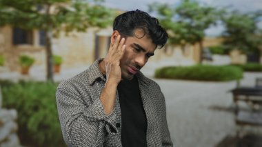 Kareli gömlekli genç adam, güneşli binanın ön cephesinde eli yanaklı duruyor; diş ağrısı şikayeti.