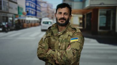 Ukrayna bayraklı askeri üniformalı bir adam açık bir şehir caddesinde güvenle duruyor..