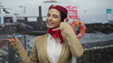 Üniformalı bir kadın sahilde kırmızı bir eşarpla el kol hareketi yapıyor, okyanus manzarasına güvenle gülümsüyor, profesyonel ama rahat bir hava katıyor, sahil zeminine yaslanıyor, seyahat teması için mükemmel..