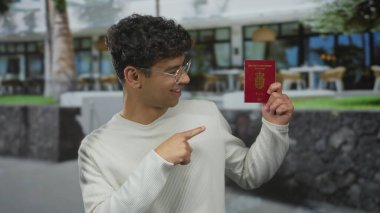 Beyaz kazak giyen bir adam dışarıdaki restoran binasında kırmızı Danimarka pasaportuna parmak işareti yapıyor; seyahat heyecanı.