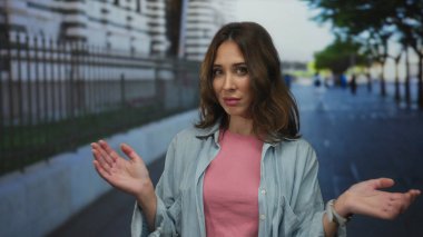 Şehir sokaklarında pembe gömlekli ve kot ceketli bir kadın var. Arka planda güneşli bir günde binalar ve ağaçlar var.