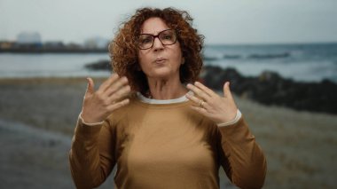 Elleriyle şehirde el kol hareketi yapan, gözlük takan, kıvırcık saçlı, İspanyol orta yaşlı, deniz kenarında yaşayan, animasyon duygusunu ve gündelik tarzını gösteren bir kadın..