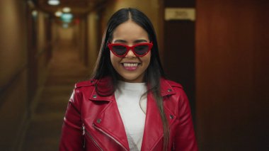 Kırmızı ceketli ve güneş gözlüklü kendine güvenen genç bir kadın loş bir otel koridorunda gülümsüyor rahat ve modern bir görünüm sergiliyor..