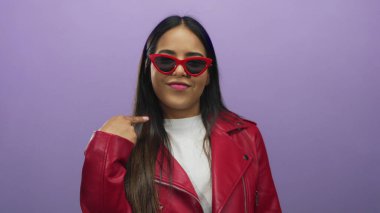 Kırmızı ceketli ve güneş gözlüklü bir kadın izole edilmiş mor arka planda, kendinden emin bir şekilde poz veriyor..