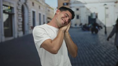Güler yüzlü İspanyol adam gündüz vakti şehir meydanındaki güneş ışığıyla aydınlanan kaldırım taşı caddesinde yanak jesti yapıyor..