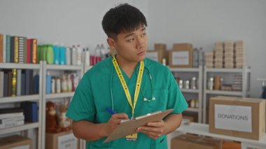 Steteskoplu yeşil önlüklü genç adam, bağış merkezinde gönüllü olarak çalışıyor, kutularla ve malzemelerle çevrili notlar yazıyor, başkalarına yardım etmeye odaklanıyor..