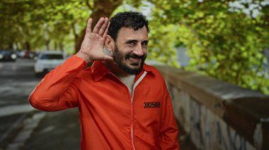 Turuncu tulumlu, sakallı bir adam caddede dikilip yeşil yaprakların ve arabaların arasında dikkatle dinliyor..